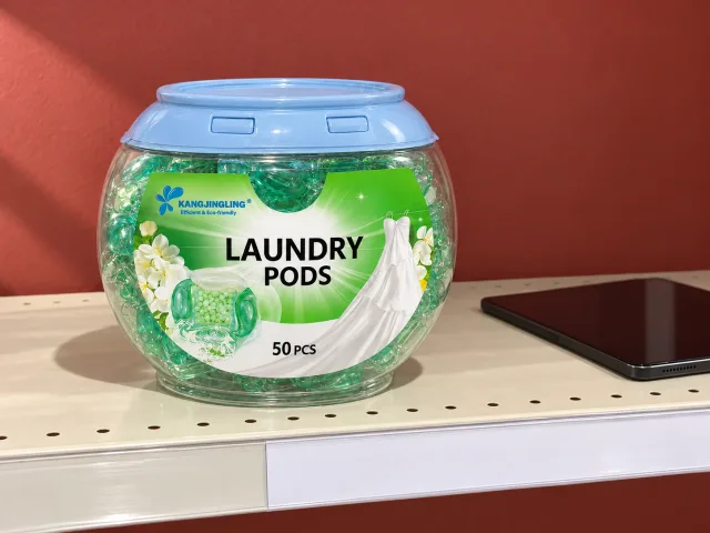 A closed Kangjingling laundry-detergent-pod tub on a neutral retail shelf beside a blank shelf ticket, in an illustrative scene.